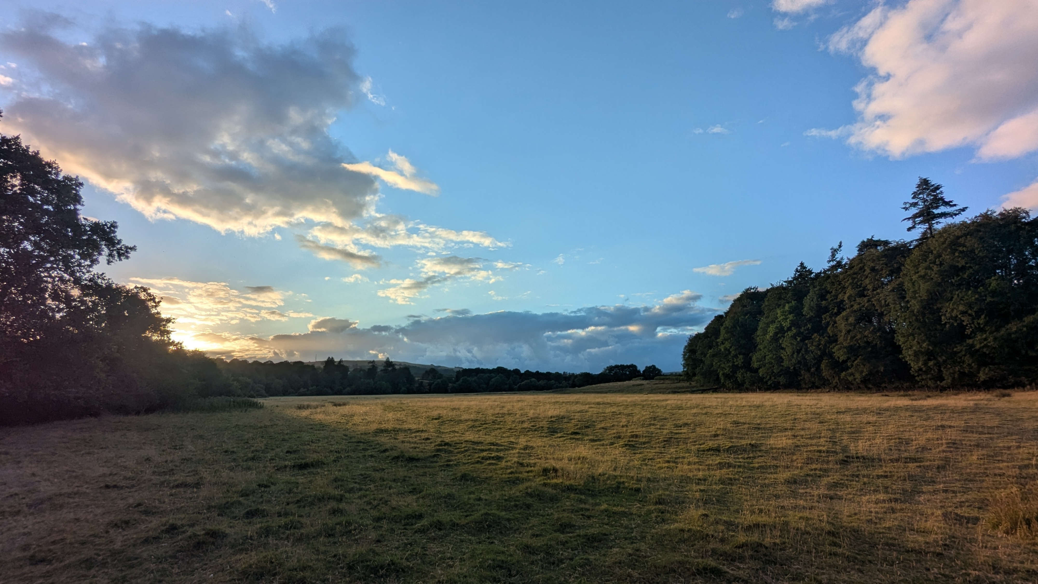 A view across a large field with trees around its border. The sun is setting in the left of frame and the blue sky is buffered by clouds. August 2026. Wales, UK.